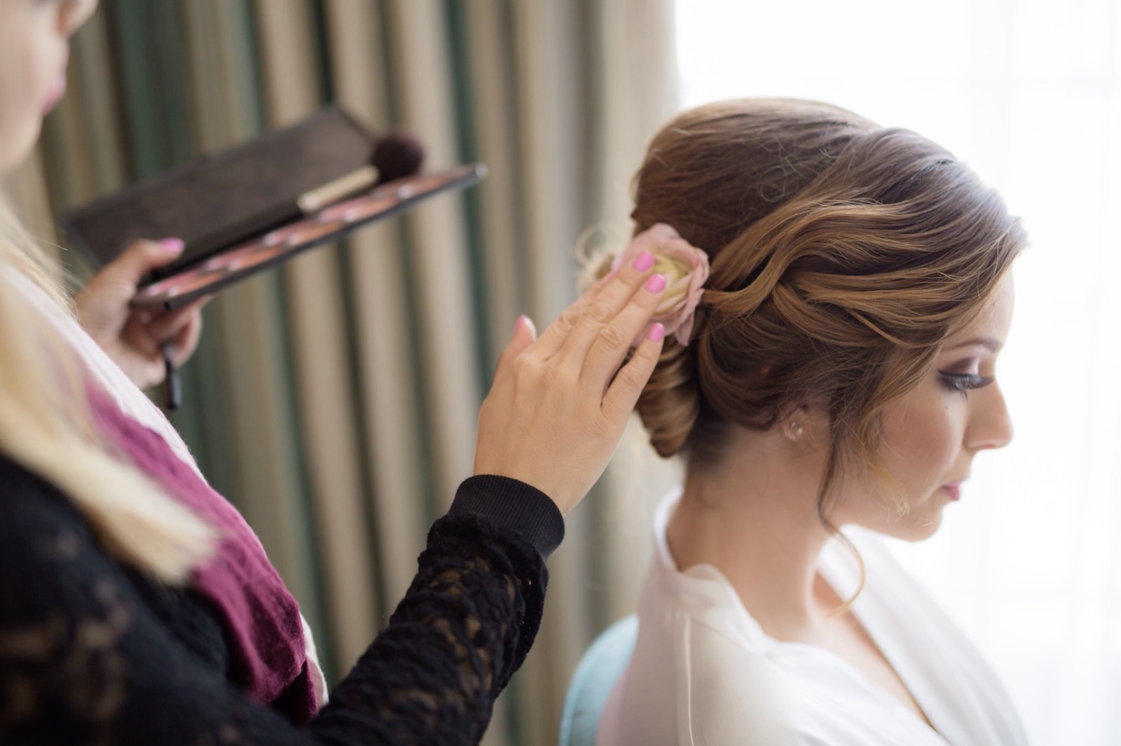 Hair stylist working on bride updo in a well-lit hotel room during prep